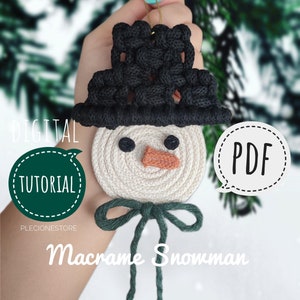 Puede incluir: Un tutorial digital en PDF para hacer un adorno de macramé en forma de muñeco de nieve. El muñeco de nieve está hecho con cordón blanco, negro y verde. El adorno tiene un sombrero negro, una cara blanca y un lazo verde.