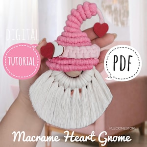 May include: A pink and white macrame heart gnome with a wooden bead nose and two red felt hearts on its hat. The gnome is hanging from a hand. The text "DIGITAL TUTORIAL" is in a pink circle with a white dotted border. The text "PDF" is in a white circle with a black dotted border. The text "Macrame Heart Gnome" is below the gnome.
