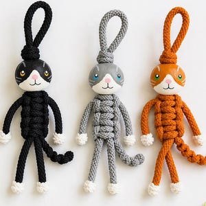 Op de afbeelding: Drie handgemaakte katvormige sleutelhangers in zwart, grijs en oranje, elk met een lus om op te hangen. De katfiguren hebben witte gezichten, poten en een staart. Een houten vis en kattenkop zijn ook aanwezig, samen met groene blad takjes.