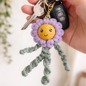 Op de afbeelding: Een handgemaakte sleutelhanger met een lachend bloemdesign. De bloem heeft een gele gezicht, paarse bloemblaadjes en groene gevlochten armen en benen. De sleutelhanger is bevestigd aan een gouden ring met sleutels en een afstandsbediening voor de auto.