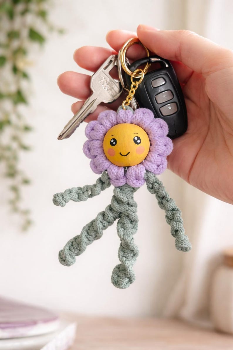 Op de afbeelding: Een handgemaakte sleutelhanger met een lachend bloemdesign. De bloem heeft een gele gezicht, paarse bloemblaadjes en groene gevlochten armen en benen. De sleutelhanger is bevestigd aan een gouden ring met sleutels en een afstandsbediening voor de auto.
