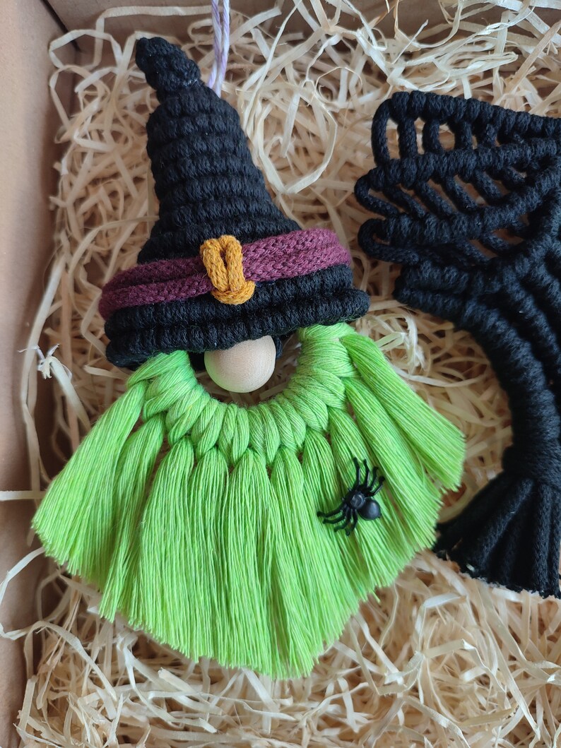 Op de afbeelding: Een groene en zwarte heks-dwerg ornament met een zwarte heksenhoed, een houten neus en een zwarte spin. De dwerg is gemaakt van garen en heeft een lange groene baard.