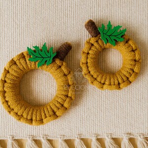 May include: Two handmade, circular pumpkin decorations. The pumpkins are made of mustard-yellow yarn, with brown stems and green leaf accents. They are placed on a cream-colored woven surface.