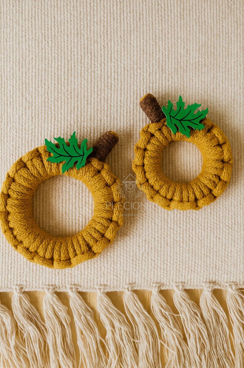May include: Two handmade, circular pumpkin decorations. The pumpkins are made of mustard-yellow yarn, with brown stems and green leaf accents. They are placed on a cream-colored woven surface.