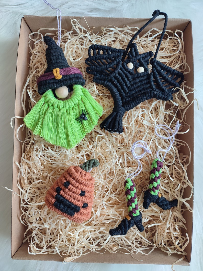 Op de afbeelding: Een set van vijf handgemaakte Halloween-versieringen in een bruine doos met gescheurd papier. De versieringen omvatten een heks, een vleermuis, een pompoen en twee heksenlaarzen. De heks is groen en heeft een zwarte hoed met een paarse band. De vleermuis is zwart en heeft een spinnenwebontwerp. De pompoen is oranje en heeft een zwart gezicht. De heksenlaarzen zijn groen en zwart.