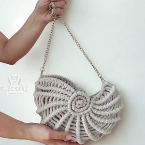 May include: A light gray macrame handbag with a silver chain strap. The bag features a spiral design in the center and a woven pattern throughout. The bag is held up by a silver chain.