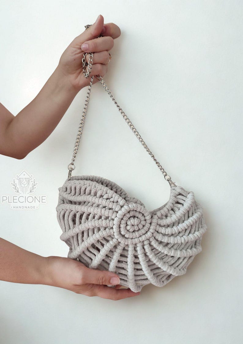 May include: A light gray macrame handbag with a silver chain strap. The bag features a spiral design in the center and a woven pattern throughout. The bag is held up by a silver chain.