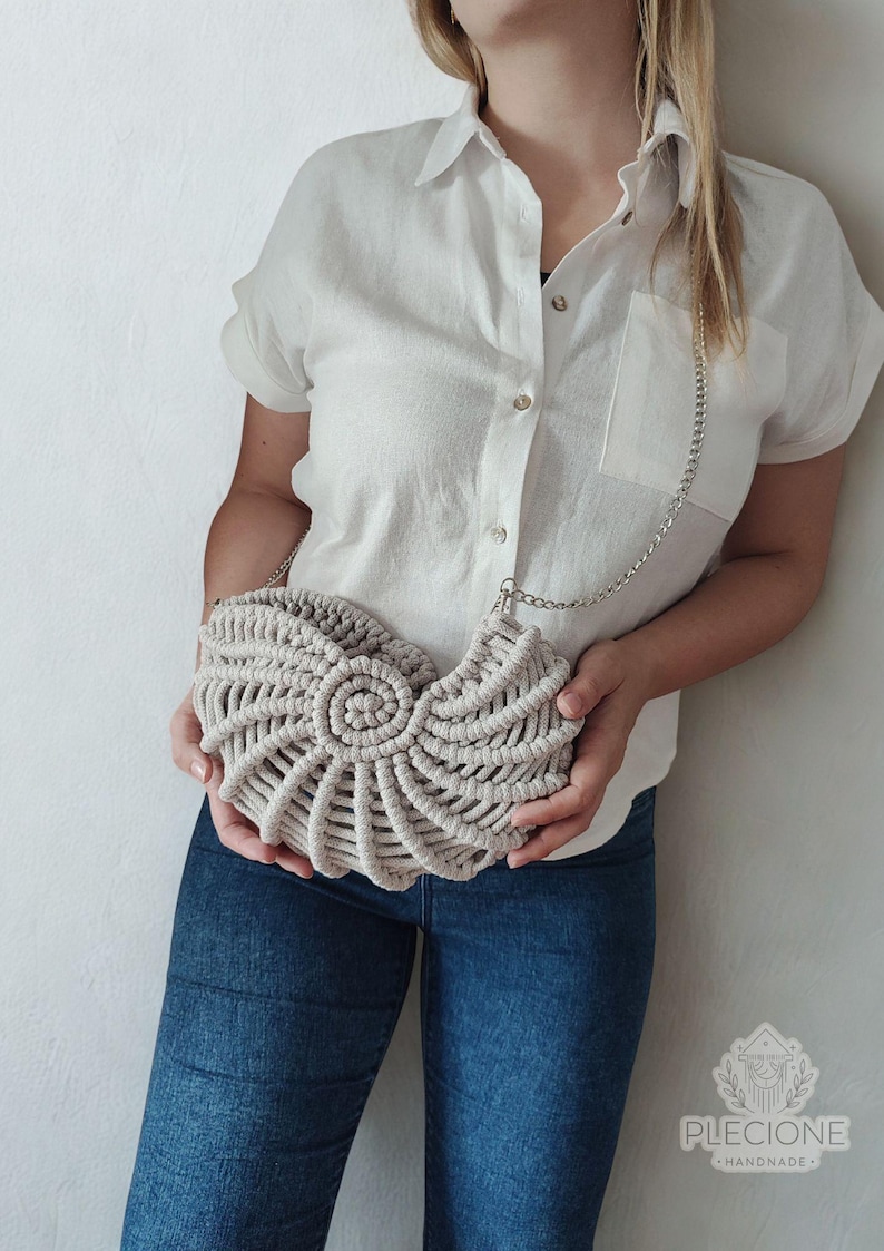 May include: A beige macrame handbag with a spiral design, featuring a silver chain strap. The bag is held against a white shirt and blue jeans. The bag's design resembles a nautilus shell.