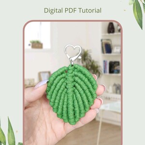 May include: A green macrame leaf keychain with a silver heart-shaped clasp. The keychain is being held in a hand. The image includes the text "Macrame Leaf" and "Digital PDF Tutorial".