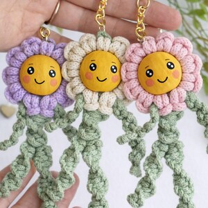 Op de afbeelding: Drie handgemaakte bloemvormige sleutelhangers in paars, cr&egrave;me en roze. Elke sleutelhanger heeft een lachend geel gezicht, een gehaakte bloemkop en groene gehaakte stelen. De sleutelhangers zijn bevestigd aan goudkleurige sleutelringen.