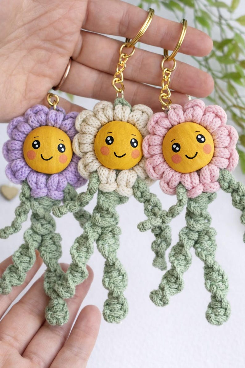 Op de afbeelding: Drie handgemaakte bloemvormige sleutelhangers in paars, cr&egrave;me en roze. Elke sleutelhanger heeft een lachend geel gezicht, een gehaakte bloemkop en groene gehaakte stelen. De sleutelhangers zijn bevestigd aan goudkleurige sleutelringen.