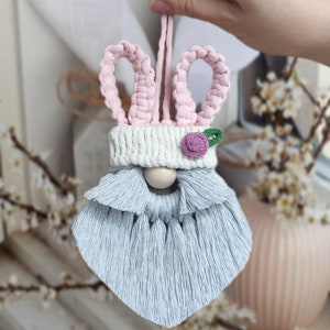 May include: A light blue and pink bunny gnome ornament with a white hat and a pink flower. The gnome has a long, fluffy beard.