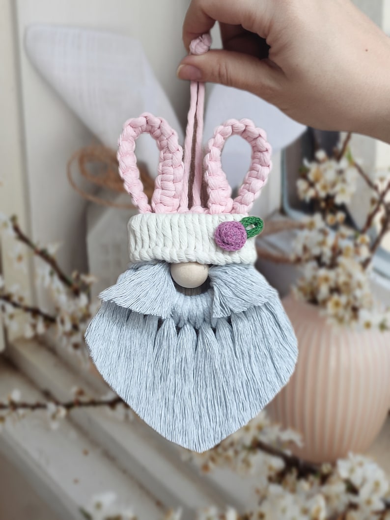 May include: A light blue and pink bunny gnome ornament with a white hat and a pink flower. The gnome has a long, fluffy beard.