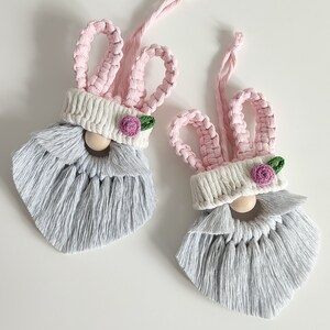 May include: Two grey and pink bunny-themed gnomes with white hats and pink flowers. The gnomes have long grey beards and are made of yarn.