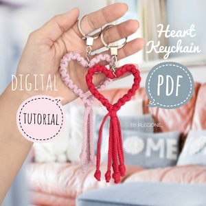 以下が含まれることがあります： ピンクと赤のロープで作ったハート型のキーホルダー2つ。タッセル付き。キーホルダーは手に持たれており、デジタルチュートリアルPDFが購入可能です。画像には「Heart Keychain」と「PDF」の文字が見えます。
