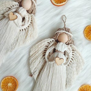 May include: Two handmade macrame angel ornaments with wooden heads and hearts. The angels are white with brown accents and are hanging from a white fuzzy surface.