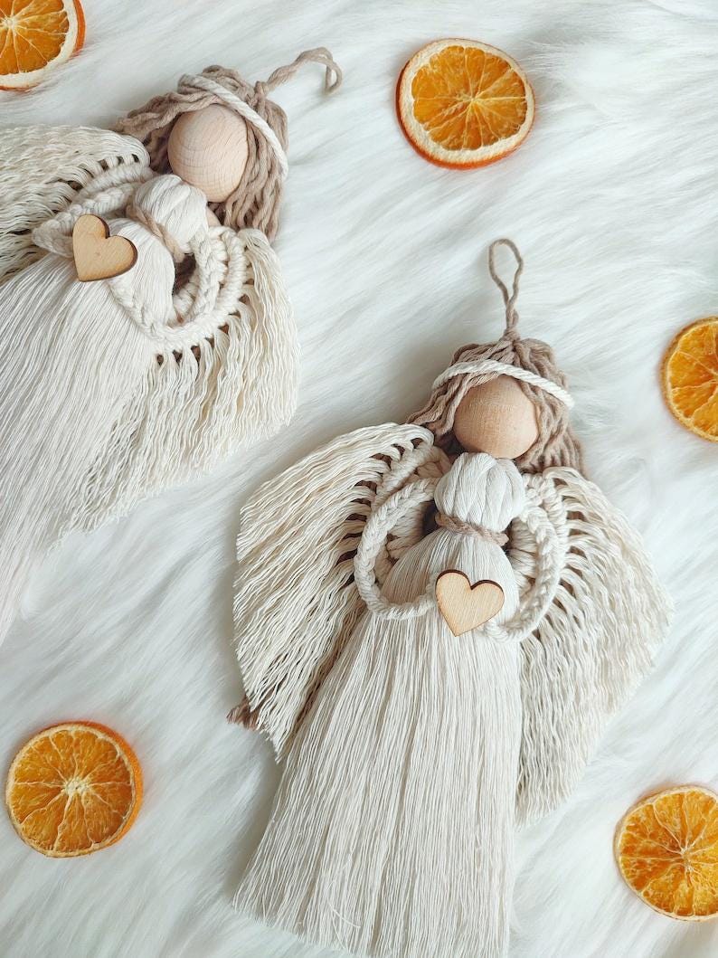 May include: Two handmade macrame angel ornaments with wooden heads and hearts. The angels are white with brown accents and are hanging from a white fuzzy surface.