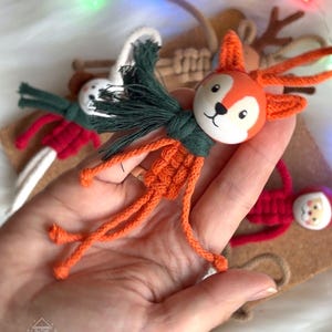 Pu&ograve; includere: Ornamento natalizio a forma di volpe arancione fatta a mano con una sciarpa verde. La volpe ha una testa di legno dipinta e un corpo in macram&egrave;. Sono visibili altre decorazioni festive, come un pupazzo di neve, Babbo Natale e renne.
