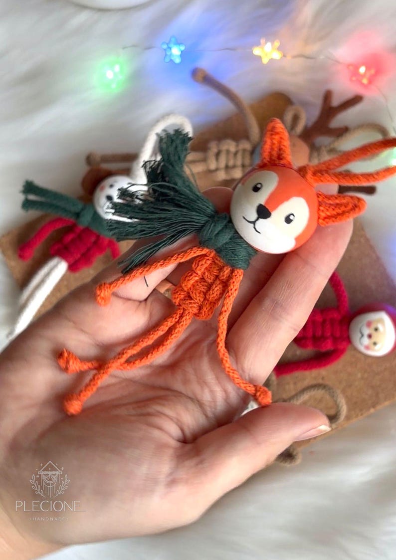 Pu&ograve; includere: Ornamento natalizio a forma di volpe arancione fatta a mano con una sciarpa verde. La volpe ha una testa di legno dipinta e un corpo in macram&egrave;. Sono visibili altre decorazioni festive, come un pupazzo di neve, Babbo Natale e renne.