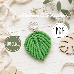 Könnte beinhalten: Ein grüner Makramee-Blatt-Schlüsselanhänger mit einem silbernen herzförmigen Verschluss. Das Bild enthält den Text Macrame leaf Keychain, DIGITAL, TUTORIAL und PDF. Das Design zeigt einen zentralen Strang mit geknoteten Kordeln in Blattform.