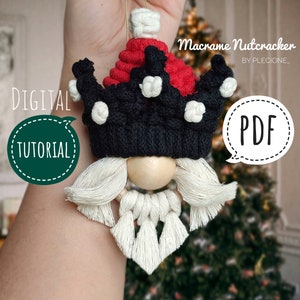May include: A macrame nutcracker ornament with a red and black crown, white beard, and wooden bead nose. The ornament is held in a hand against a blurred background of a Christmas tree. The text "Macrame Nutcracker" and "PDF" are visible in the image.