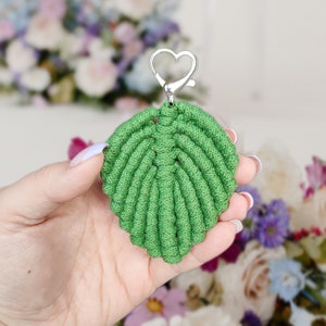 May include: A green leaf-shaped keychain made with macrame knotting. The keychain has a silver heart-shaped clasp.
