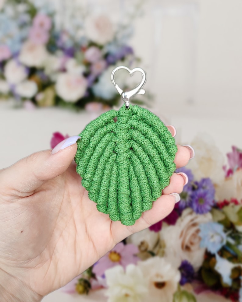 May include: A green leaf-shaped keychain made with macrame knotting. The keychain has a silver heart-shaped clasp.