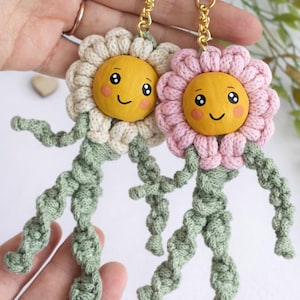 May include: Two handmade flower keychains. One has a cream petal design, the other a pink petal design. Both have yellow faces with smiling expressions and green crocheted stems and legs. The keychains are attached to gold-coloured key rings.