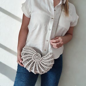 May include: A beige macrame handbag with a silver chain strap. The bag has a seashell design and is held by a person wearing a white button-down shirt and blue jeans. The bag is handmade.