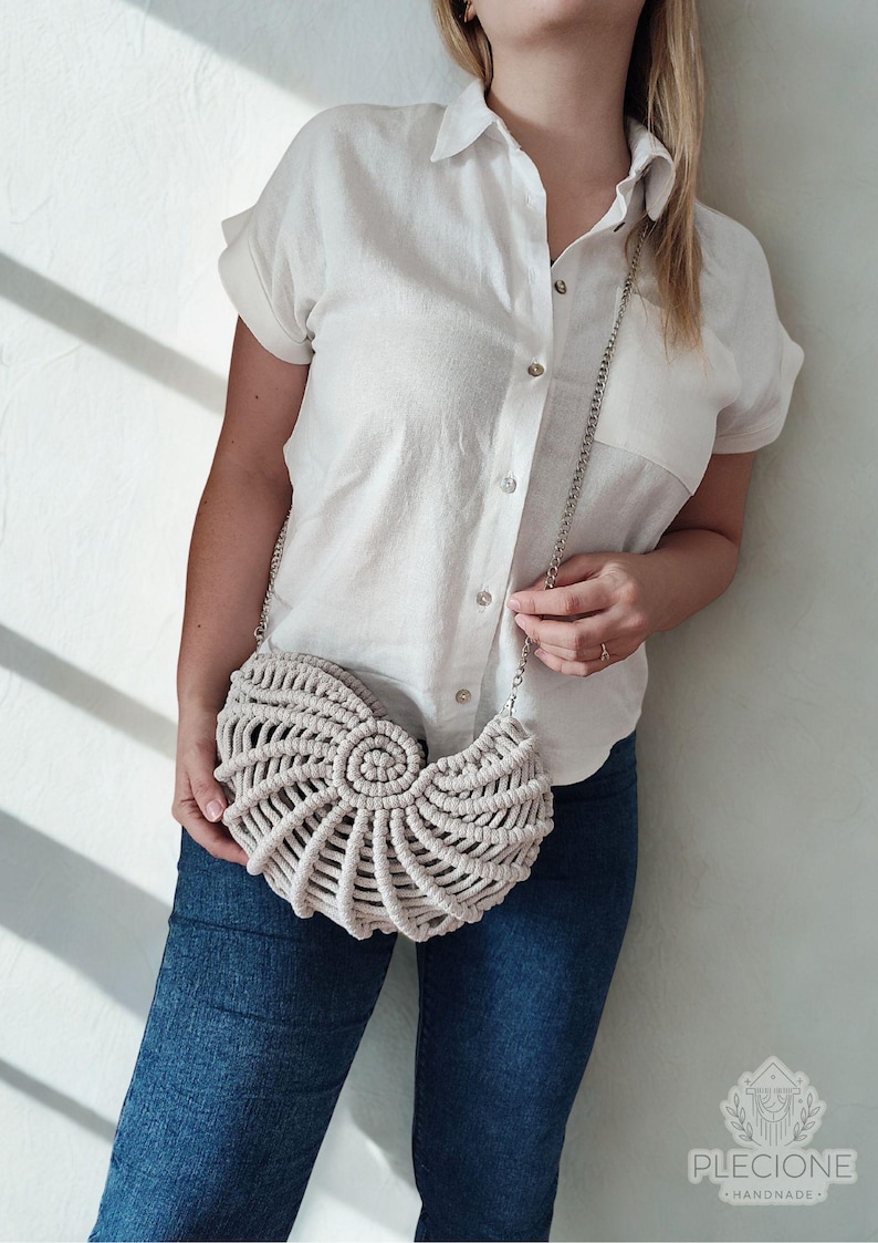 May include: A beige macrame handbag with a silver chain strap. The bag has a seashell design and is held by a person wearing a white button-down shirt and blue jeans. The bag is handmade.