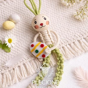 May include: A handmade Easter bunny figure with a cream body, green braided legs and ears, and a painted face. The bunny holds a felt egg decorated with red, yellow, and green dots. The background includes decorative eggs and a white fringed blanket.
