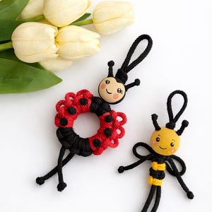 Puede incluir: Dos llaveros artesanales con tem&aacute;tica de insectos. Uno es una mariquita con un dise&ntilde;o rojo y negro, y el otro es una abeja con rayas amarillas y negras. Ambos tienen caras sonrientes y detalles de cord&oacute;n negro. Hay tulipanes blancos en el fondo.