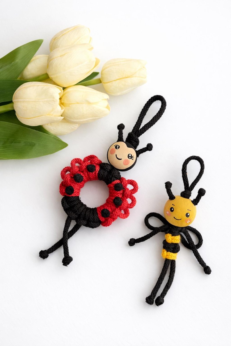 Puede incluir: Dos llaveros artesanales con tem&aacute;tica de insectos. Uno es una mariquita con un dise&ntilde;o rojo y negro, y el otro es una abeja con rayas amarillas y negras. Ambos tienen caras sonrientes y detalles de cord&oacute;n negro. Hay tulipanes blancos en el fondo.