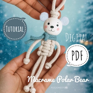 May include: A handmade macrame polar bear keychain with a white body, light blue scarf, and small white ears. The image includes the text "Tutorial" and "Digital PDF".