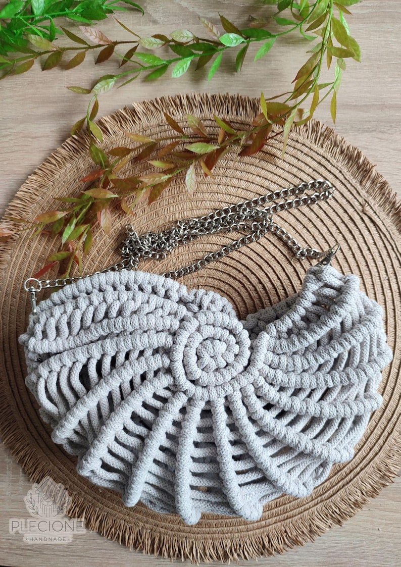 May include: A light gray macrame handbag with a silver chain strap. The bag features a spiral design in the center and a woven pattern. The bag is displayed on a woven mat with green and brown leaves.