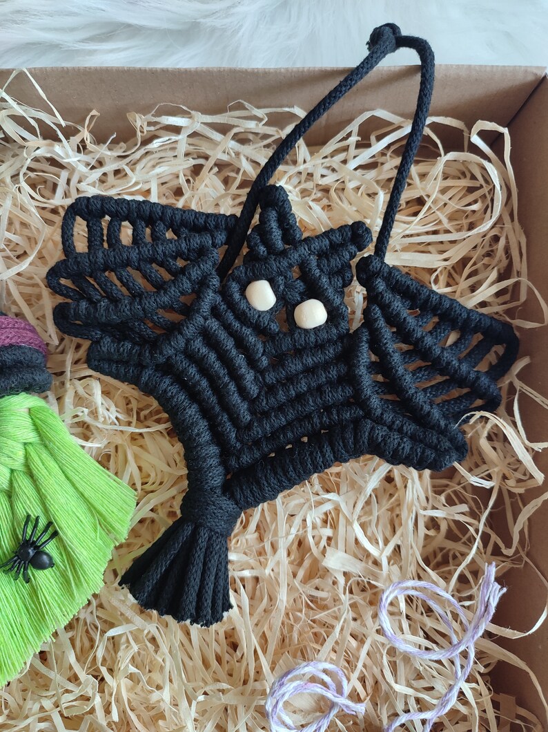 Op de afbeelding: Zwarte macram&eacute; vleermuis wanddecoratie met witte houten kralen voor de ogen. De vleermuis hangt in een doos gevuld met gescheurd papier.