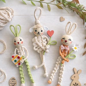 May include: Three handmade Easter bunny and lamb ornaments. The bunny figures have wooden heads, macrame bodies, and green or orange accents. One holds a decorated egg, another a flower, and the third a heart. Decorative wooden shapes and greenery surround them.