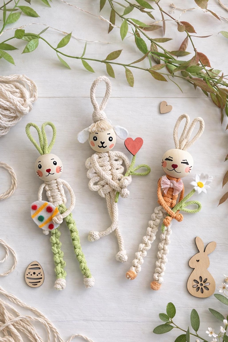 May include: Three handmade Easter bunny and lamb ornaments. The bunny figures have wooden heads, macrame bodies, and green or orange accents. One holds a decorated egg, another a flower, and the third a heart. Decorative wooden shapes and greenery surround them.