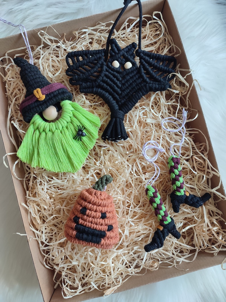 Op de afbeelding: Een set van vijf handgemaakte macram&eacute; Halloween-versieringen. De versieringen omvatten een heks, een vleermuis, een pompoen en twee heksenbenen. De versieringen zijn gemaakt van zwart, groen, oranje en bruin garen.