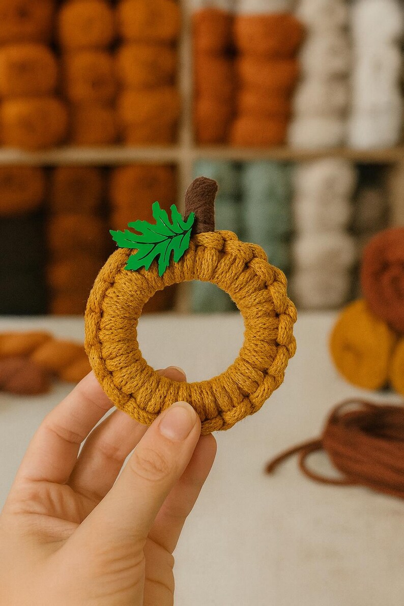 May include: A handmade, circular pumpkin decoration in a warm mustard yellow color. The pumpkin has a brown stem and a green leaf accent. The background features spools of yarn in various fall colors.