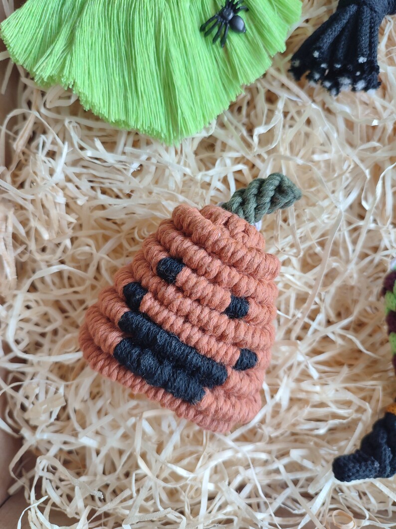 Op de afbeelding: Een klein oranje en zwart pompoen-vormig ornament gemaakt van garen. Het ornament heeft een groene steel en ligt op een bed van versnipperd papier.
