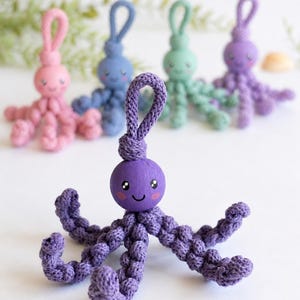 Puede incluir: Una colecci&oacute;n de llaveros de pulpo hechos a mano en varios colores pastel. Cada llavero presenta una cabeza de madera con una cara sonriente pintada y tent&aacute;culos tejidos. Los llaveros son de color rosa, azul, verde y morado.