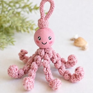 Puede incluir: Un juguete de pulpo rosa tejido a ganchillo con una cara sonriente y ocho tent&aacute;culos. El pulpo tiene un lazo en la parte superior para colgar. El fondo es blanco con un toque de vegetaci&oacute;n.