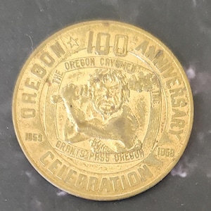 May include: A gold-colored coin commemorating the 100th Anniversary of the Oregon Cavemen Inc. The coin features an image of a caveman and the text "Grants Pass Oregon Celebration 1959".