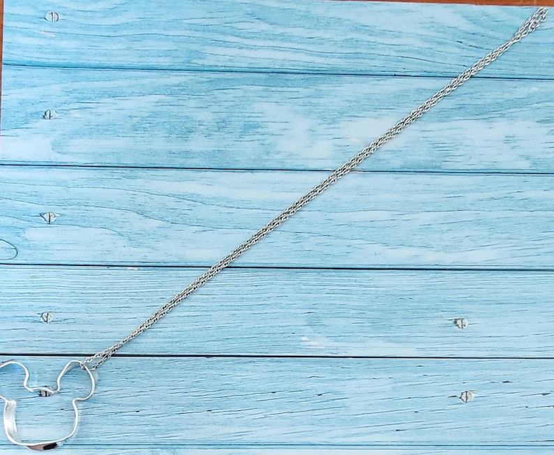 Puede incluir: Un collar plateado con un colgante de la cabeza de Mickey Mouse. El colgante es un contorno simple de la cara del personaje. La cadena tiene un dise&ntilde;o tejido. El collar se exhibe sobre una superficie de madera azul claro.