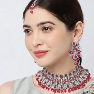 Puede incluir: Un conjunto de joyas de novia plateado y rosa con un collar choker, pendientes y un maang tikka. El collar presenta un diseño en capas con piedras rosas y detalles blancos. Los pendientes son largos y colgantes con piedras rosas y detalles blancos. El maang tikka es un tocado tradicional indio con piedras rosas y detalles blancos.