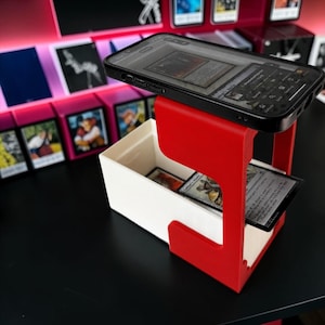 Peut inclure: Un support en plastique rouge maintenant un smartphone noir sur le dessus, positionné au-dessus d'une boîte de rangement rectangulaire blanche contenant plusieurs cartes à collectionner. L'écran affiche une interface numérique.