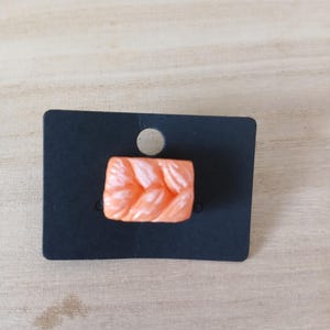 May include: A small, rectangular, peach-coloured charm featuring a raised, braided texture. The charm is attached to a black, rectangular display card with a circular hole at the top centre for hanging, designed for retail presentation.