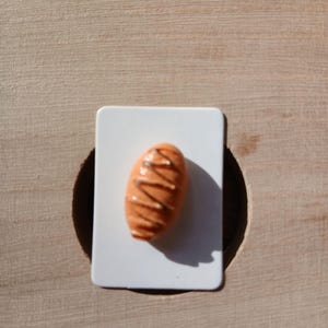 May include: A small, oval-shaped orange pastry figurine with brown drizzled icing, mounted on a rectangular white card. The miniature baked good features a textured surface and a glossy finish, casting a distinct shadow on the white backing.