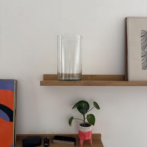 Op de afbeelding: Een heldere, cilindrische glazen vaas staat op een houten plank. Daaronder een kleine potplant met groene bladeren en een rood-wit gestreepte pot. Andere decoratieve items staan ook op de plank.